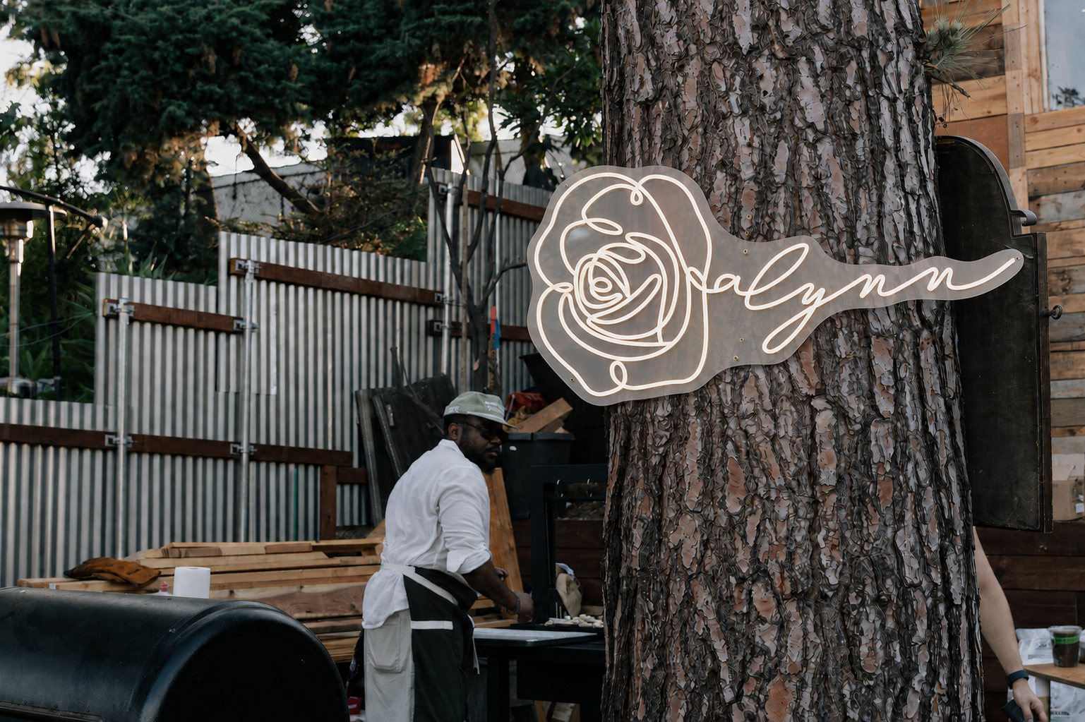 The illuminated Rosalynn rose sign mounted on a pine at Hidden Farm LA, with a chef at the prep table behind